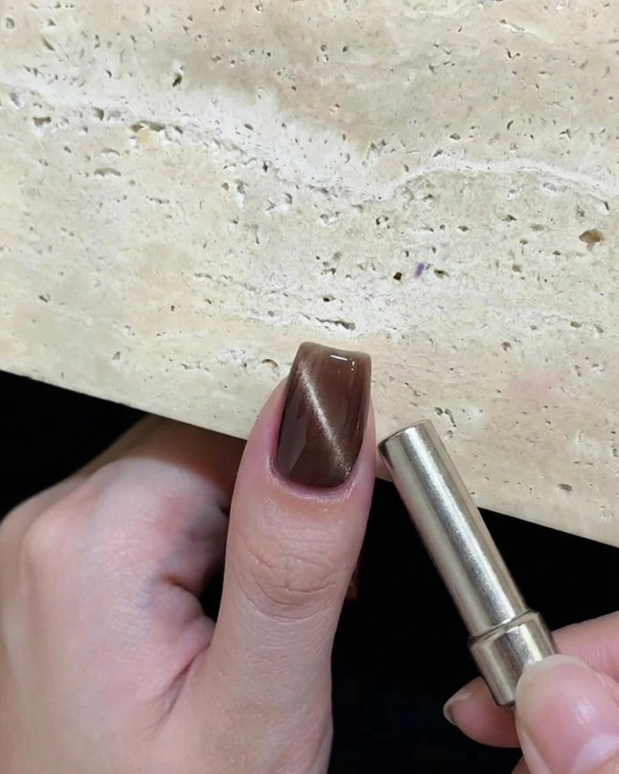 Eine Hand hält einen zylindrischen Magneten nahe an einen feuchten braunen Cat-Eye-Gelnagel, um ein kreisförmiges reflektierendes Muster zu erzeugen.