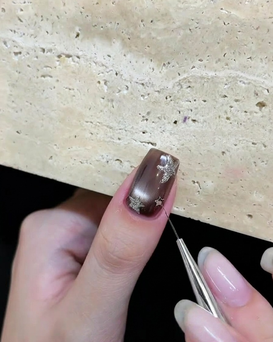 Ein Werkzeug platziert silberne Stern-Glitzerstücke auf einem braunen Gelnagel.