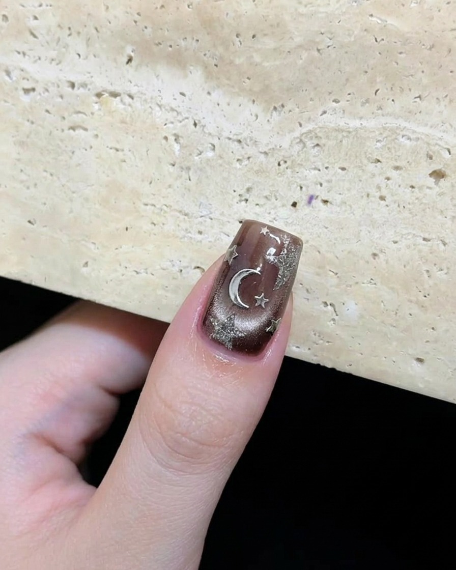 Nahaufnahme eines fertigen dunkelbraunen Nagels mit silbernen Sternen und einem künstlerisch angeordneten Mond-Akzent.