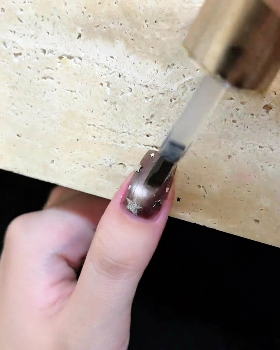 Ein Pinsel trägt einen klaren, glänzenden Top Coat über einen dunkelbraunen Nagel mit silbernen Sternen und Monden auf.