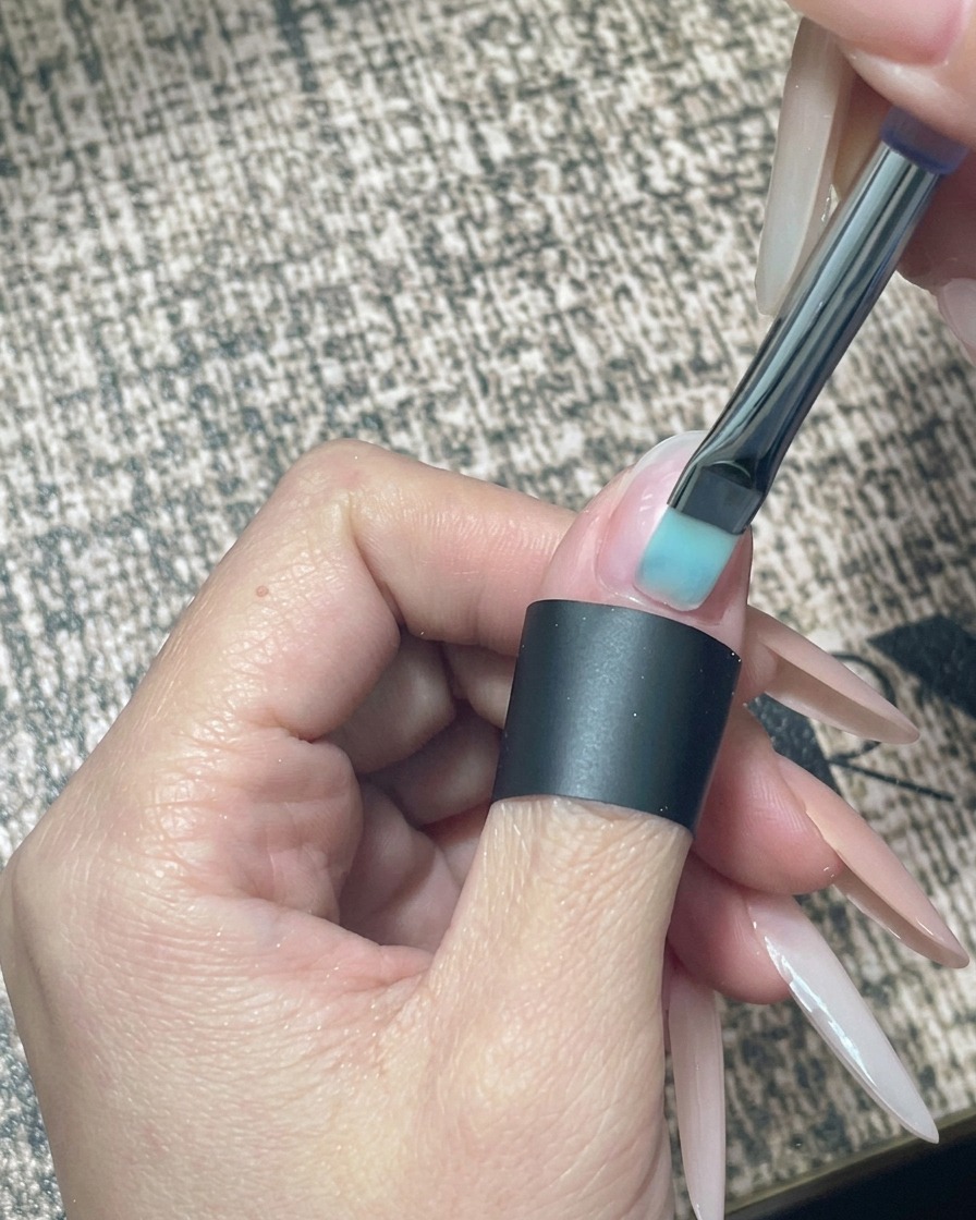 Nahaufnahme eines Pinsels, der einen transparenten Nude-Gellack auf einen vorbereiteten Naturnagel aufträgt.