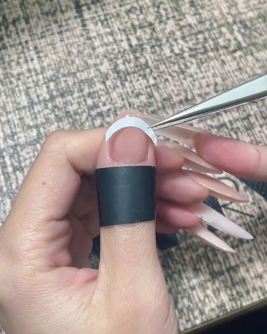 Eine Pinzette platziert einen weißen, gebogenen French-Sticker horizontal über der Mitte des Nagels.