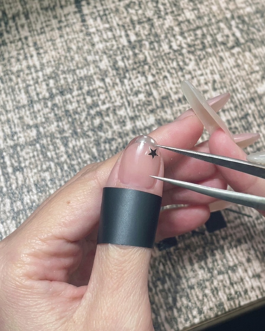 Eine Hand hält eine Pinzette, um einen kleinen schwarzen Stern-Sticker auf einen Nude-Gellack-Nagel zu platzieren.