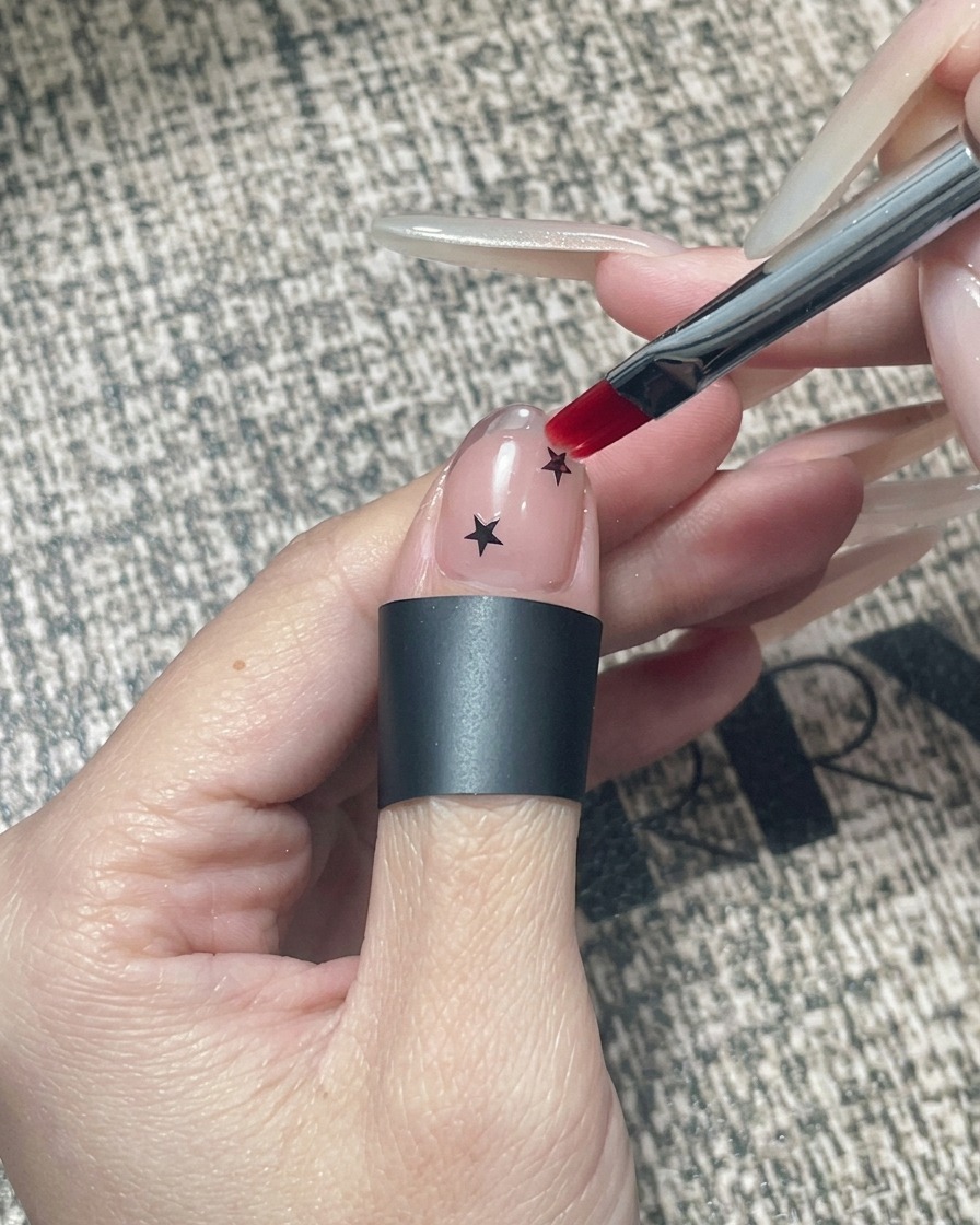 Ein quadratischer Gelpinsel trägt klares Versiegelungsgel über einen Nagel mit Sternen und Chrom-Spitze auf.