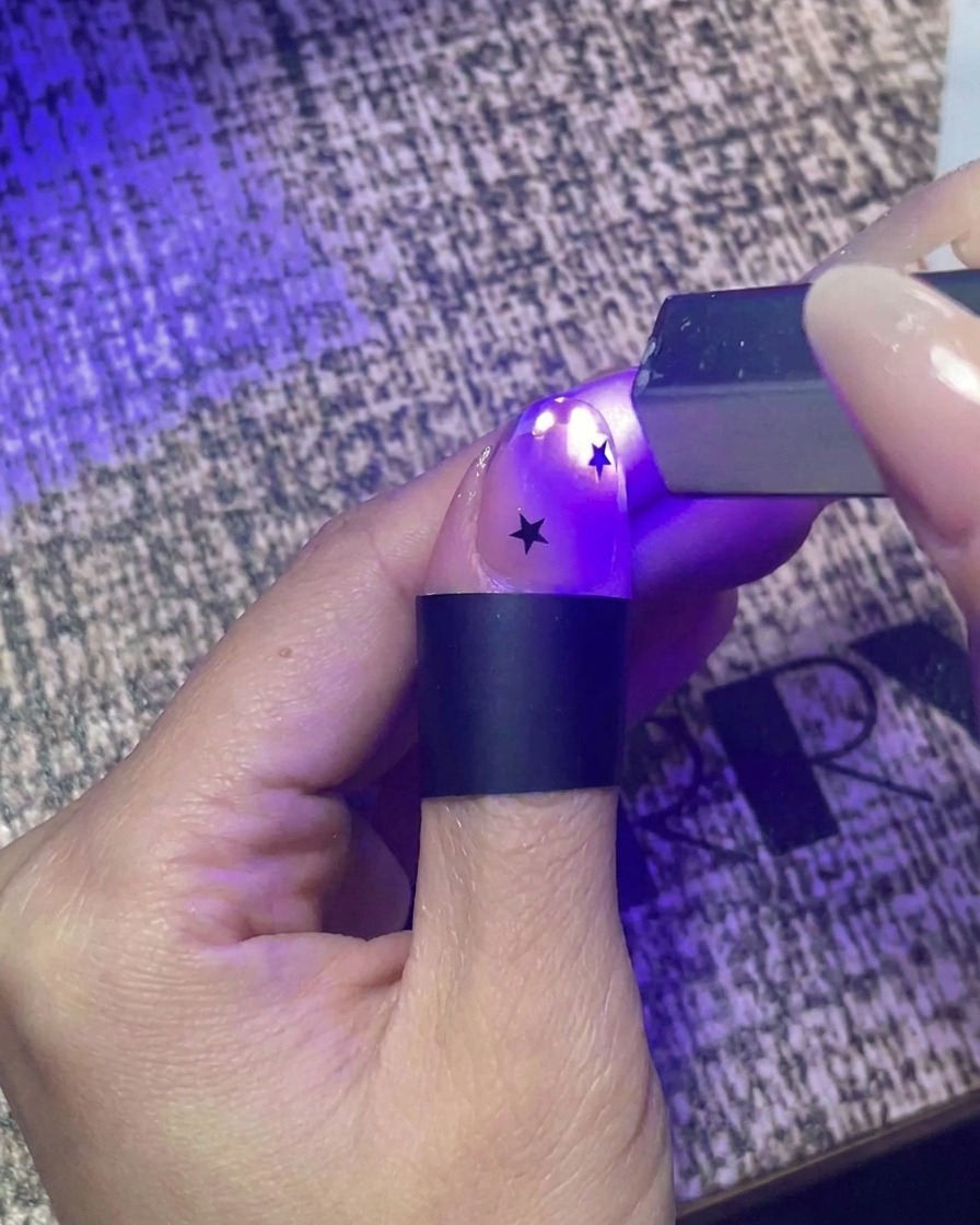 Nahaufnahme einer Hand, die eine kleine UV-Taschenlampe benutzt, um einen mit Sternen verzierten Nagel kurz auszuhärten.