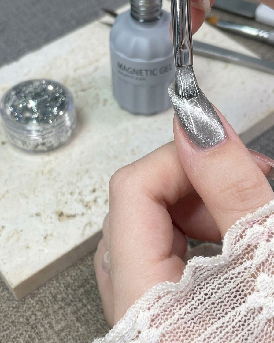Ein Pinsel trägt eine Schicht silbernes Cat-Eye-Gel auf einen natürlichen Fingernagel auf.