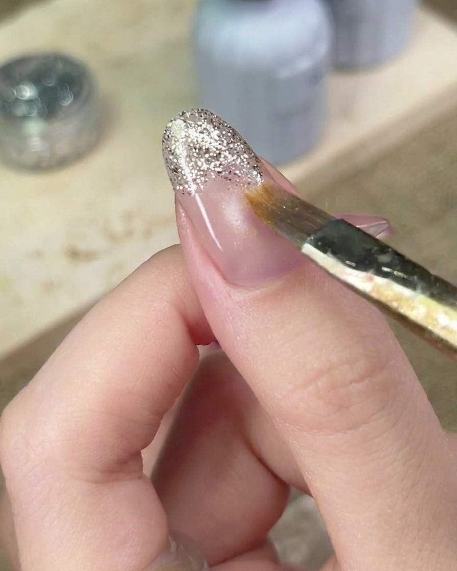 Ein Nail-Art-Pinsel verblendet sorgfältig die Ränder des silbernen Glitzer-Gels auf dem Nagel, um den Verlauf zu perfektionieren.