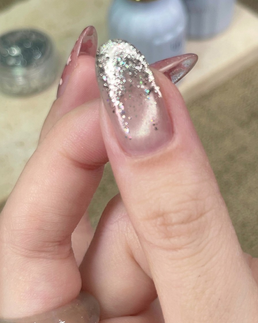 Nahaufnahme eines Werkzeugs, das eine kleine, metallische Stern-Applikation auf einen Nagel mit Silber-Glitzer-Verlauf platziert.