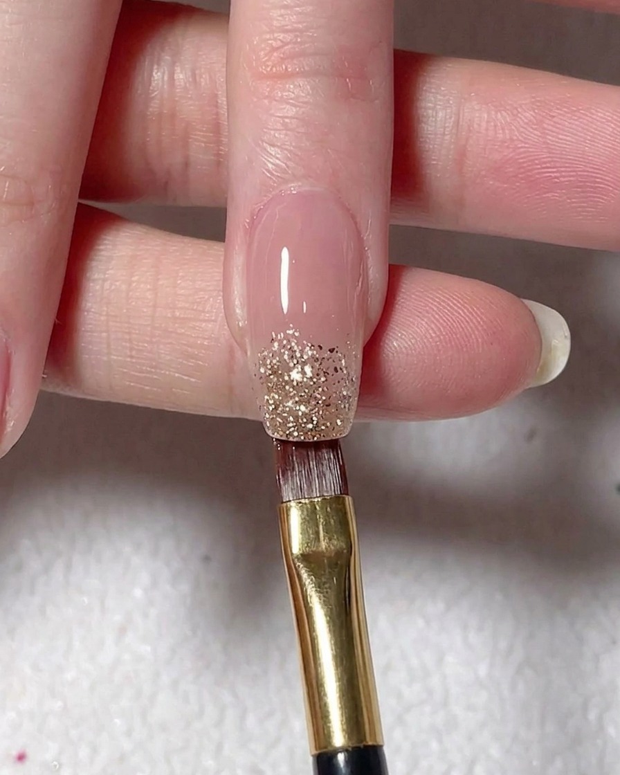 Ein Flachpinsel platziert silbernes Glitzergel auf der Spitze eines Nude-lackierten Nagels.