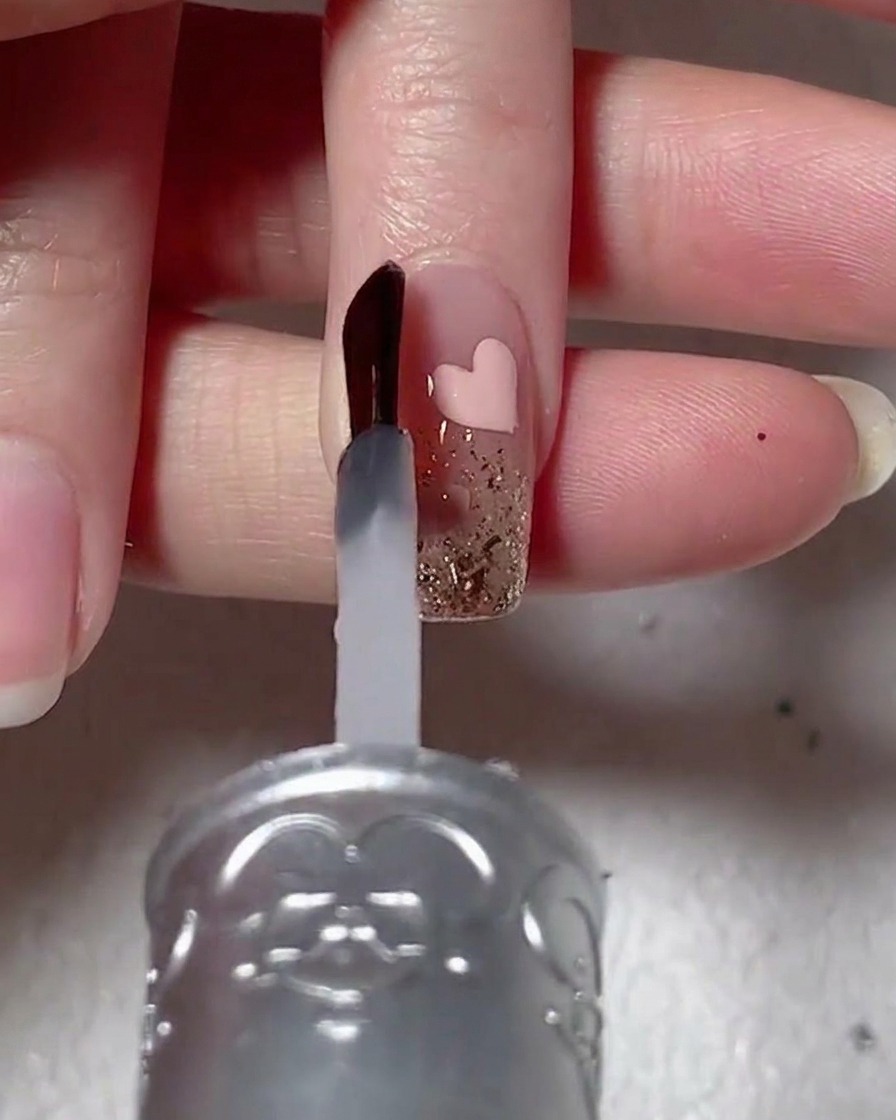 Ein Nagelpinsel trägt klares, viskoses Aufbaugel auf einen Nagel mit Glitzer und pinkem Herz-Design auf.