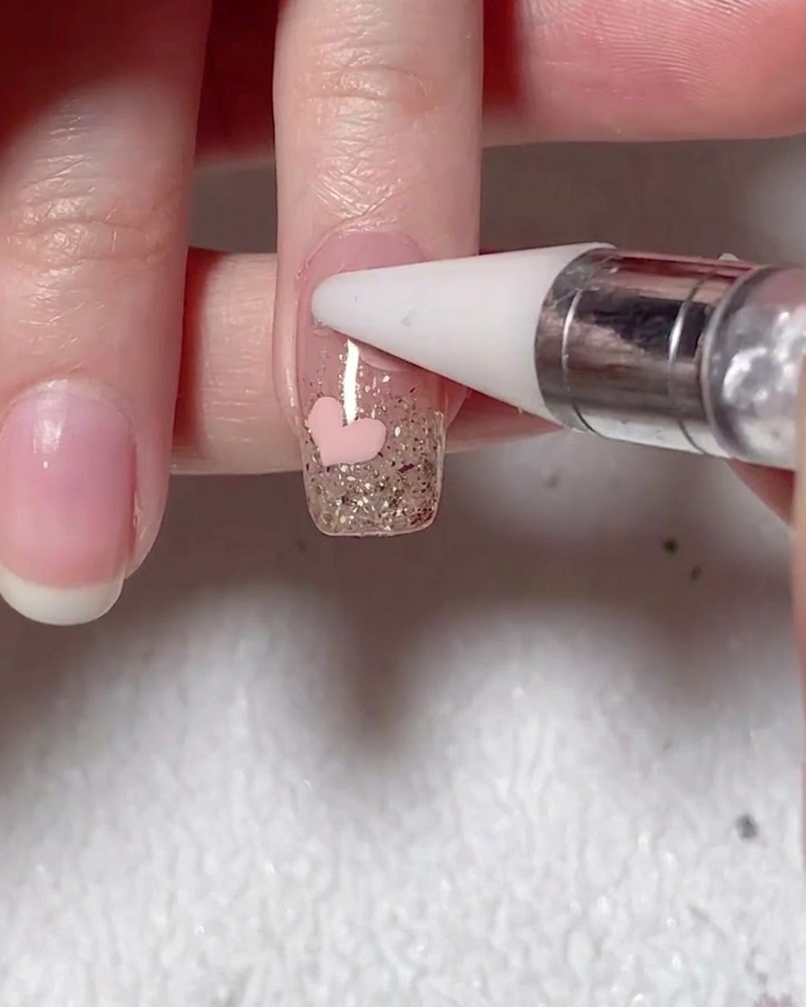 Ein Wachsstift platziert eine kleine Perle auf der Nageloberfläche neben einem gemalten pinken Herz.
