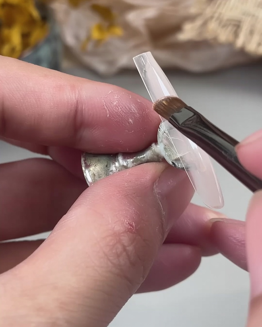 Ein Nagelpinsel trägt eine milchige, nudefarbene Gel-Basis auf einen klaren Nageltip auf, der von Fingern gehalten wird.