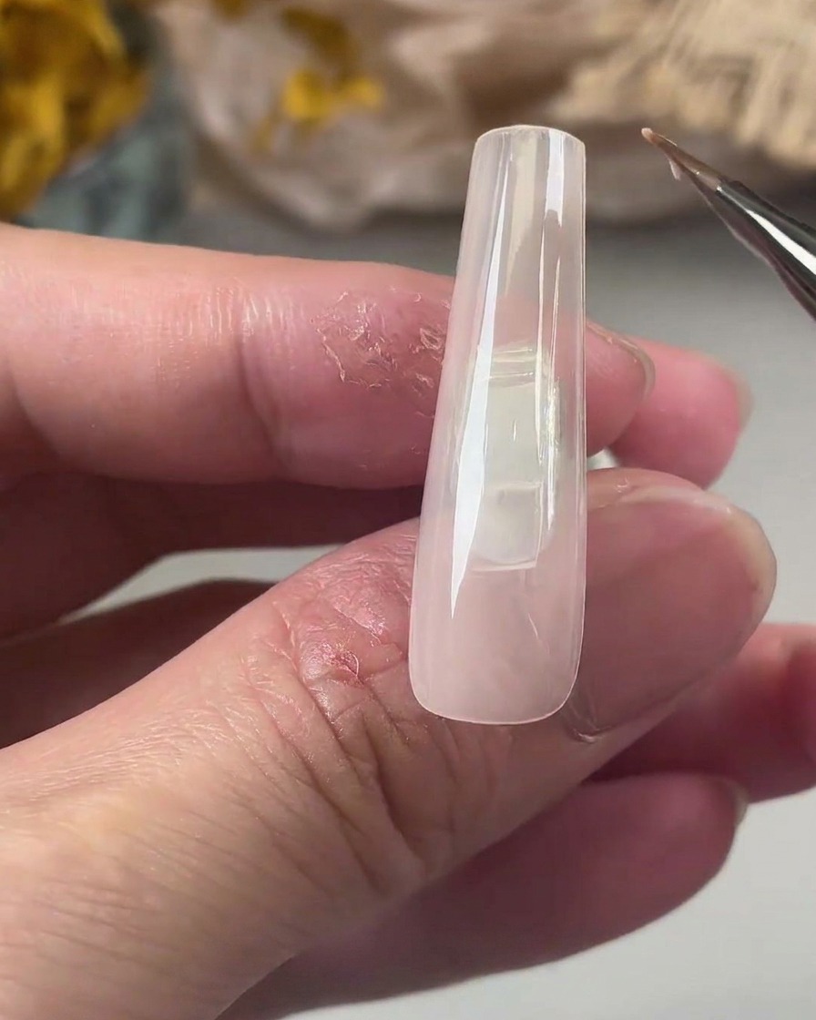 Ein feiner Detailpinsel trägt eine gleichmäßige Schicht Base Coat Gel auf einen langen, nudefarbenen, quadratischen Nageltip auf.