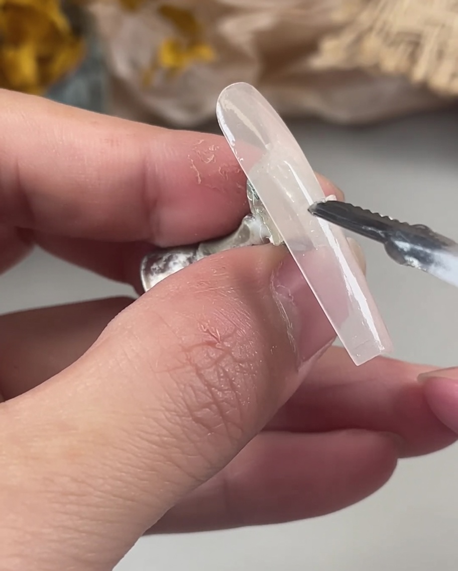 Eine Hand verwendet ein Werkzeug, um irisierende, holografische Glitzerflocken auf das nasse, ungehärtete Gel eines Nageltips zu setzen.