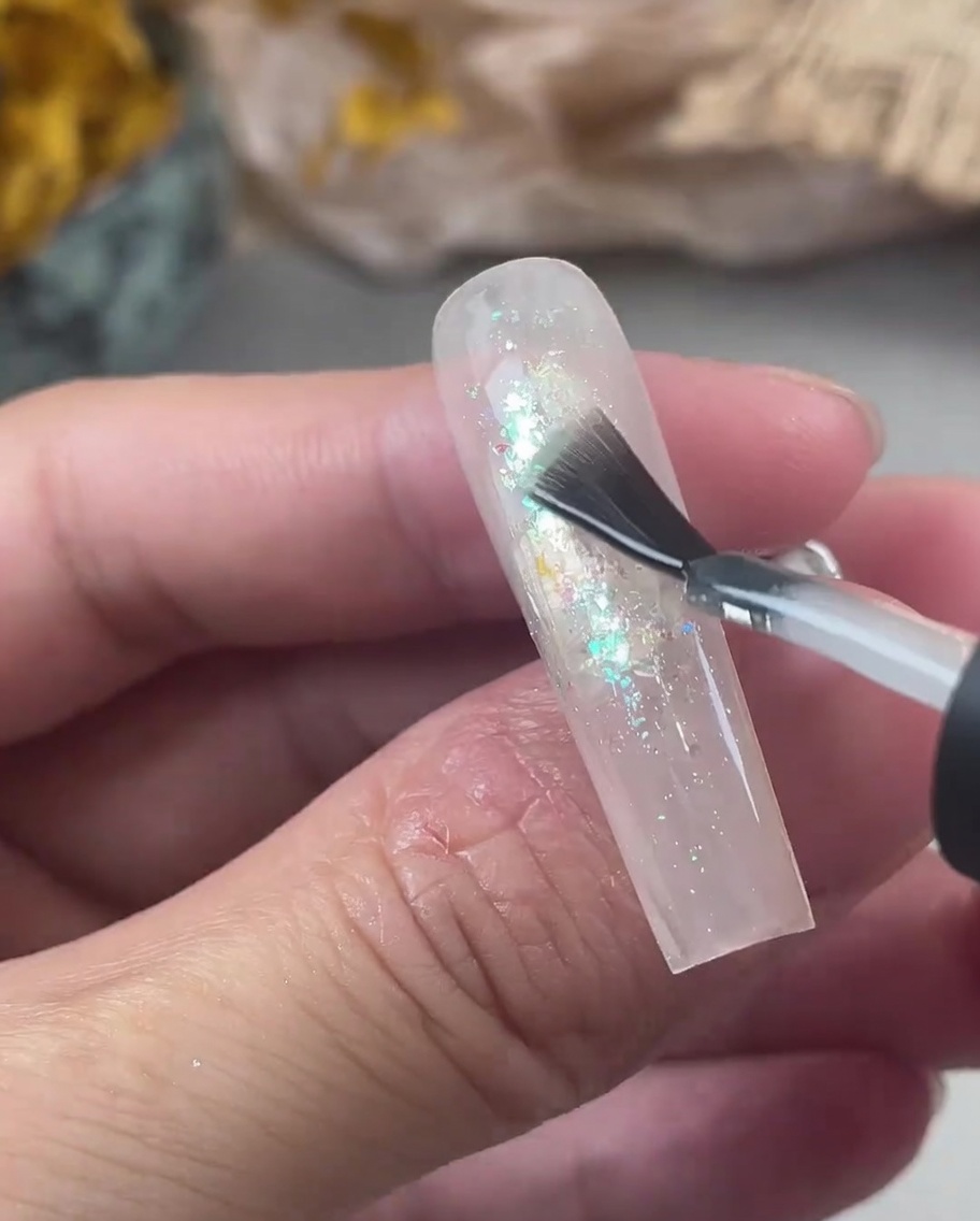 Ein Nagelpinsel trägt eine dicke Schicht klares Aufbaugel über einen Nagel mit irisierendem Glitzer auf.