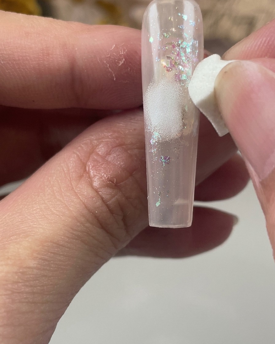 Ein weißer Kosmetikschwamm tupft sanft einen weichen Fleck weißer Farbe auf die Mitte eines Nagels mit Glitzer-Encapsulation.