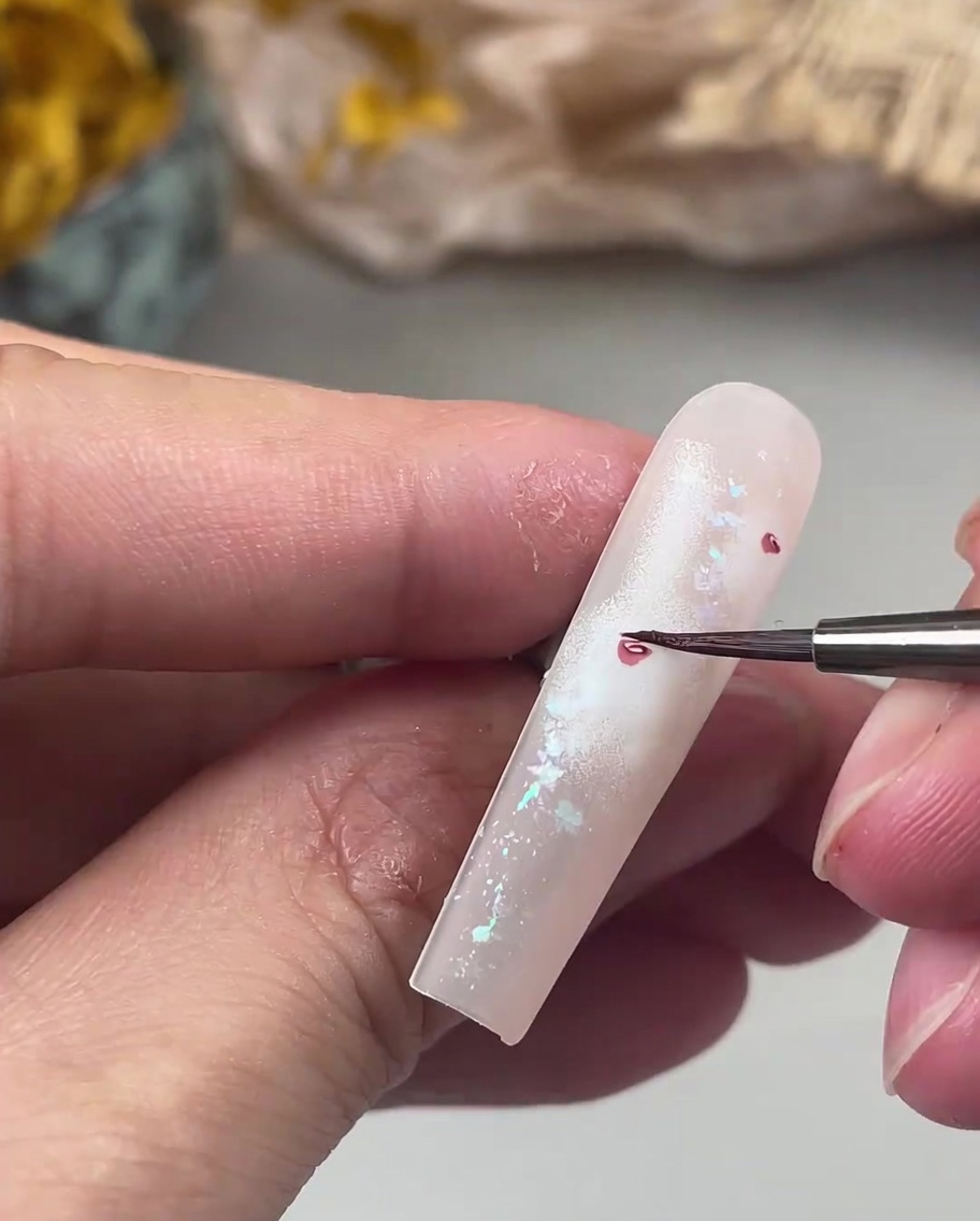 Ein feiner Nail-Art-Detailpinsel malt zarte, kleine dunkelpinke Blütenblätter um ein zentrales Rosenherz auf einem Nagel.