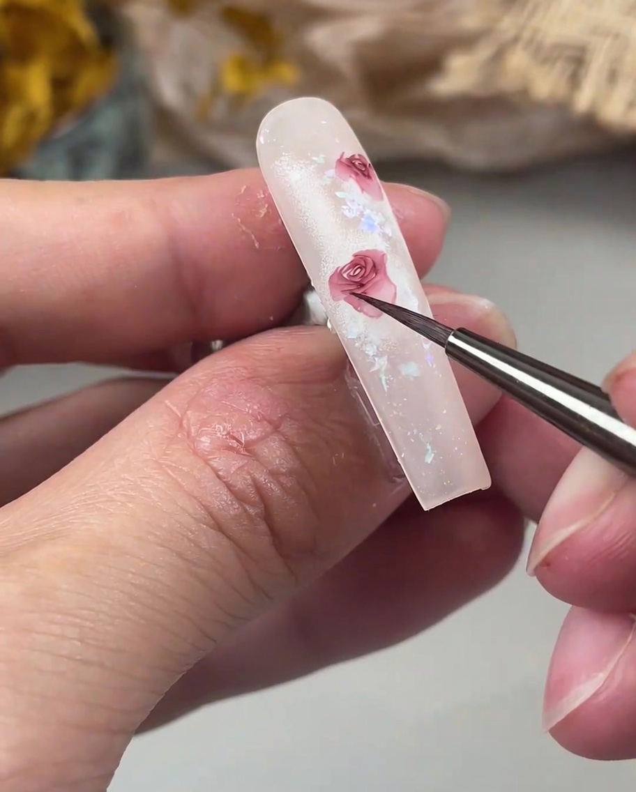 Ein Detailpinsel fügt zarte, nach außen geschwungene Blütenblätter an den Rand eines Rosendesigns auf einem Nagel hinzu.
