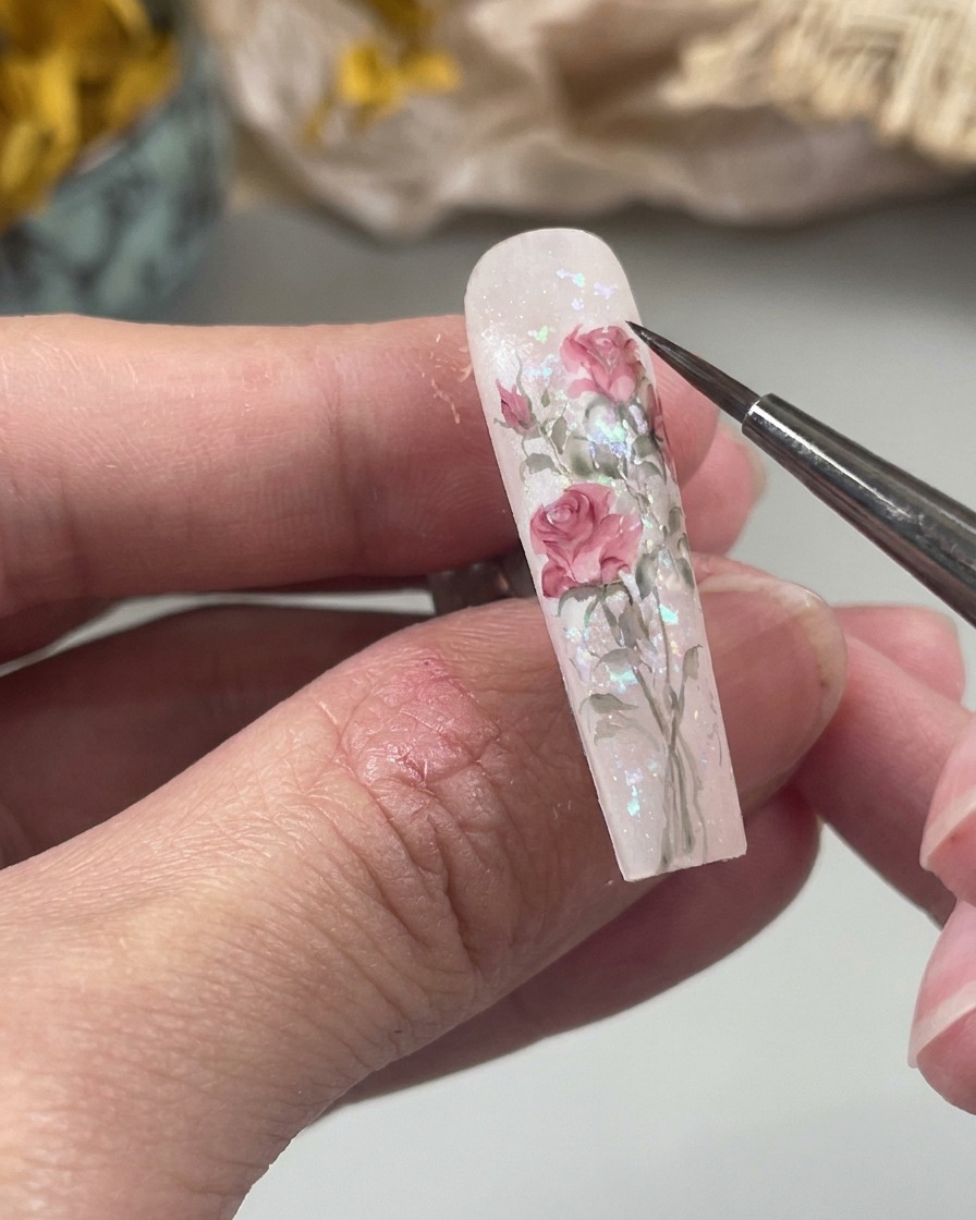 Eine Nahaufnahme eines feinen Nail-Art-Pinsels, der eine kleine, weiche pinke Wassertropfenform auf eine Nageloberfläche als Teil eines floralen Designs malt.