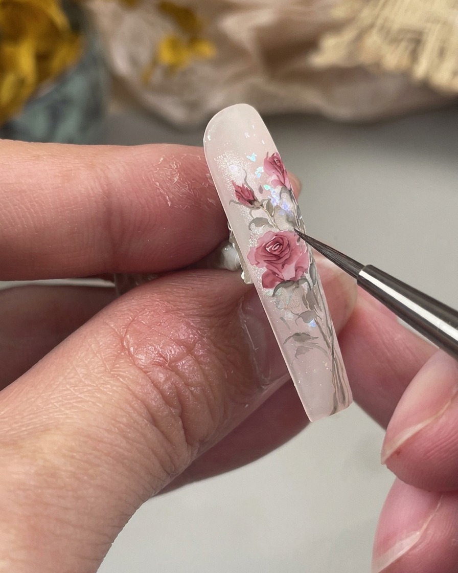 Eine Nahaufnahme zeigt einen feinen Pinsel, der kleine, spitz zulaufende grüne Blätter an der Basis einer rosenfarbenen Blumenmalerei auf dem Nagel malt.