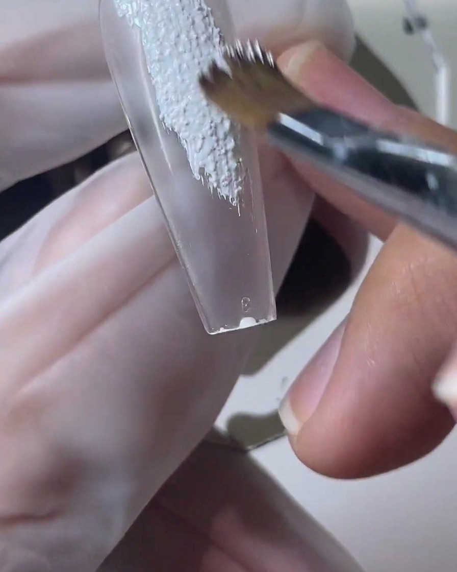 Ein Pinsel trägt strukturiertes weißes Sandgel auf einen klaren Nageltip auf.