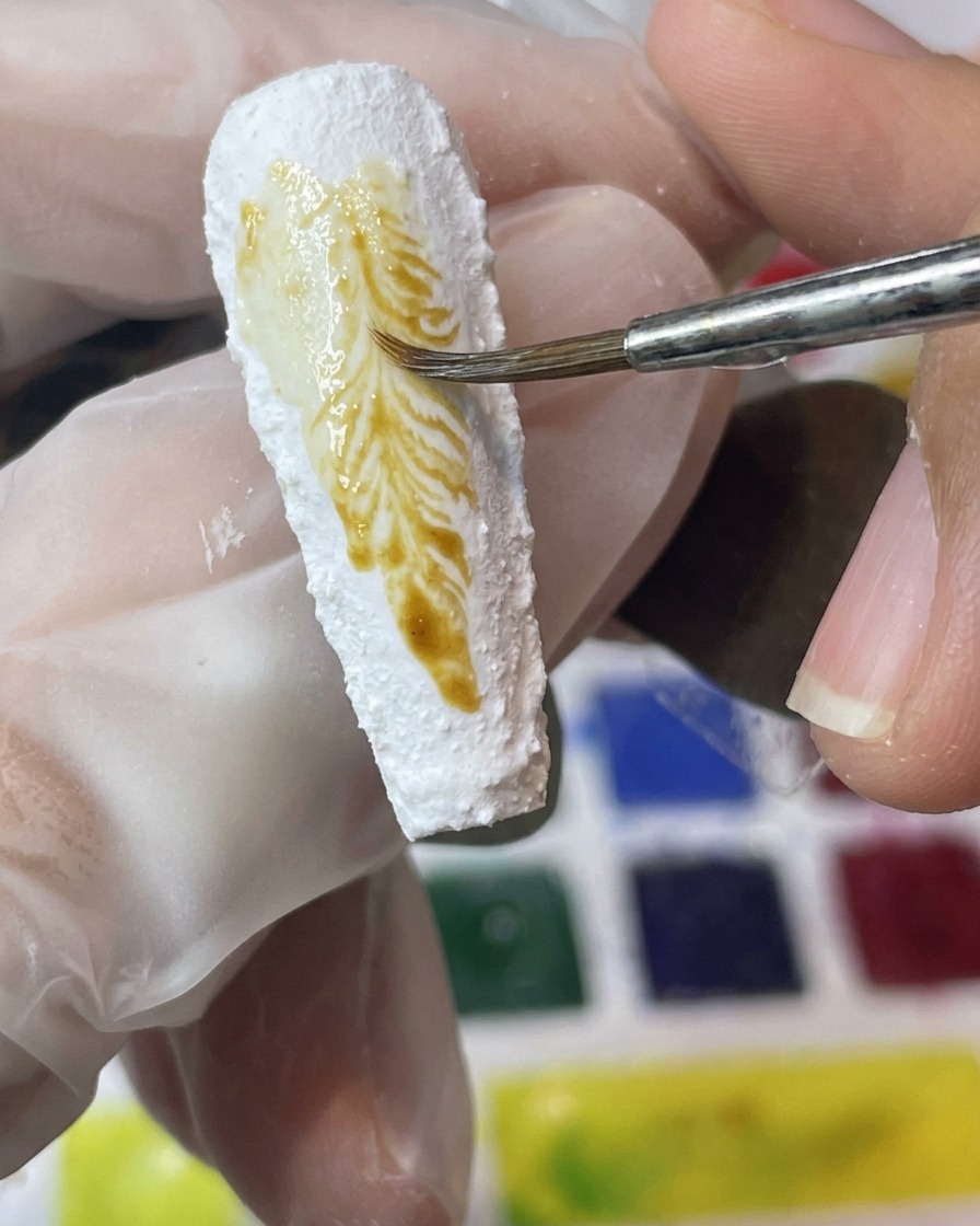 Ein Pinsel appliziert gelbe und braune Pigmente auf eine strukturierte weiße Nageloberfläche.