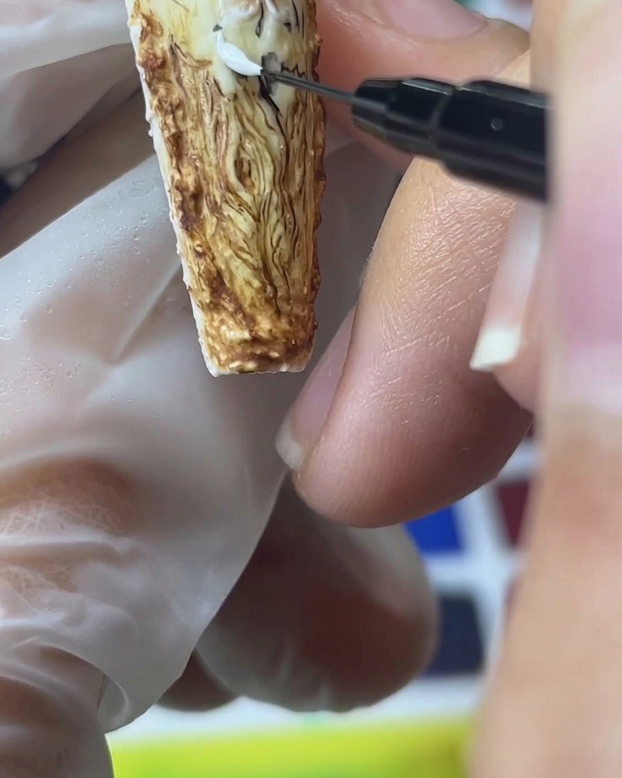 Nahaufnahme eines feinen Detailpinsels, der weißes Sandgel aufträgt, um strukturierte, plastische Rosenblätter auf braunem Grund zu formen.