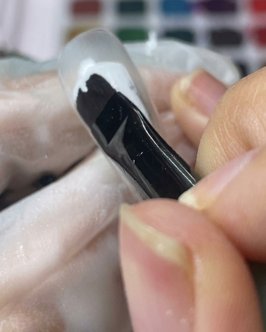 Ein schwarzer Gelpinsel trägt eine glatte Schicht weißes Gel auf einen klaren Nageltip auf.