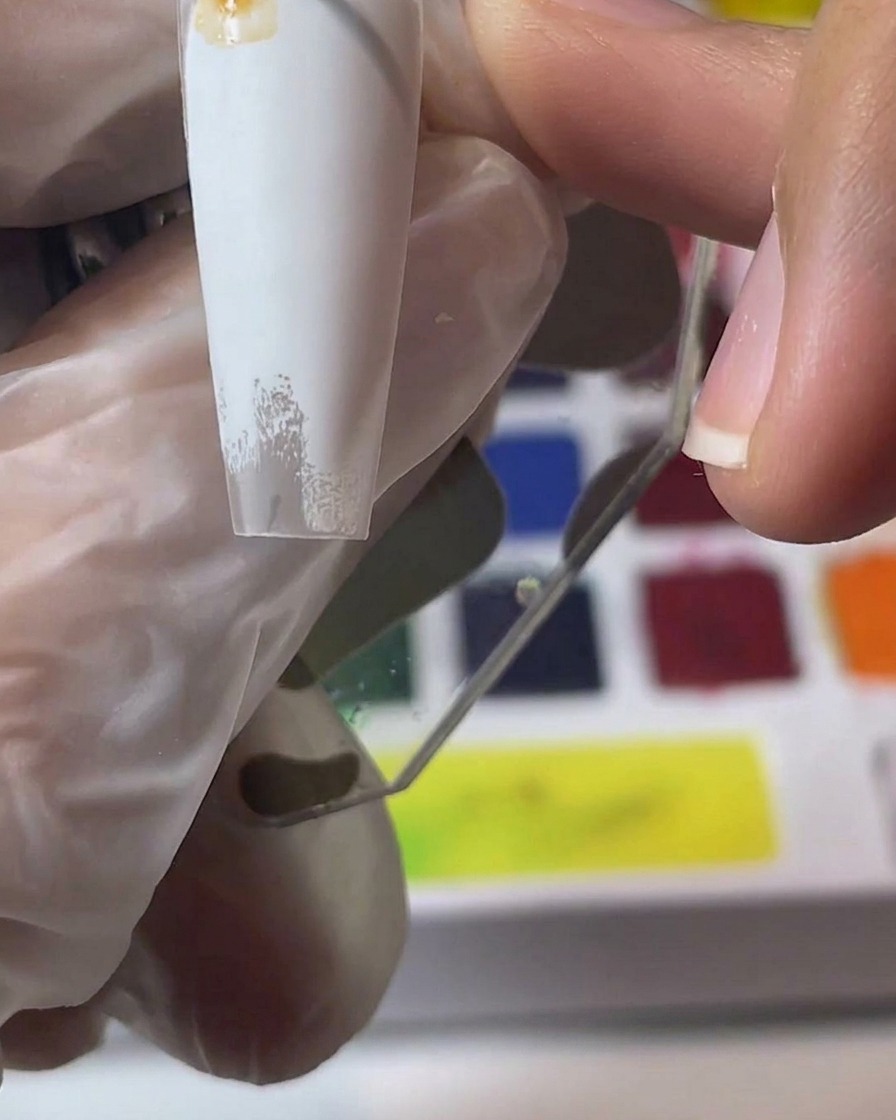 Eine Hand hält einen weißen Nageltip, während braune Aquarellpigmente gemischt und aufgetragen werden.