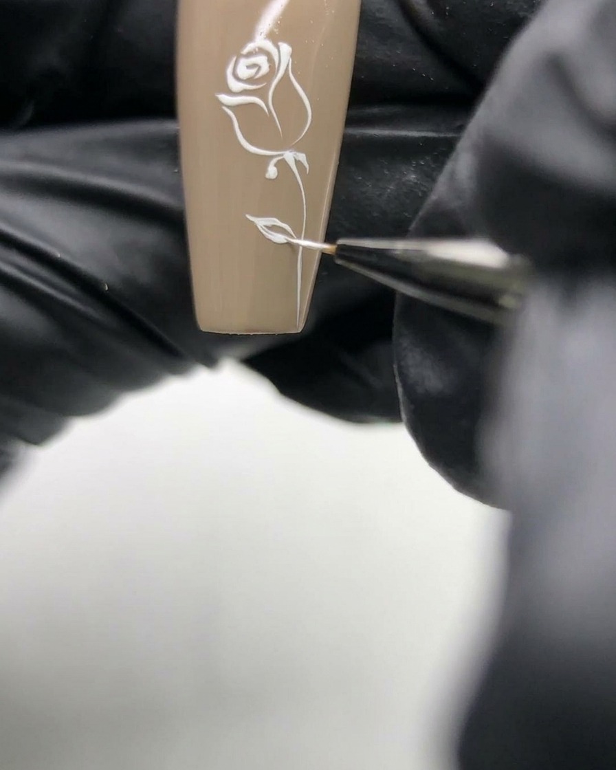 Makroaufnahme einer Nailartist, die die zarten weißen Linien eines Rosendesigns auf einem neutralen Nagel verfeinert.