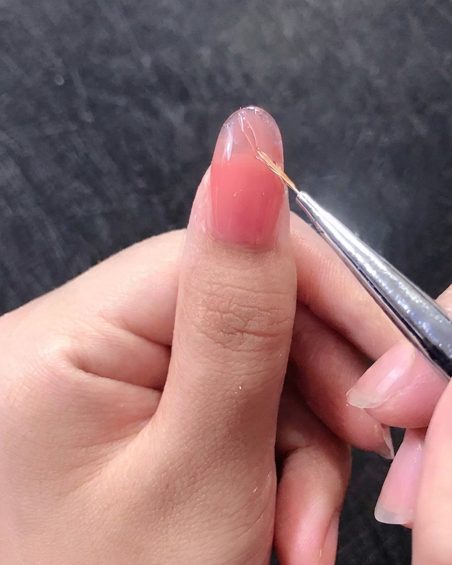 Nahaufnahme eines Nagelpinsels, der feine goldene Linien auf einen rosafarbenen Gelnagel zeichnet.