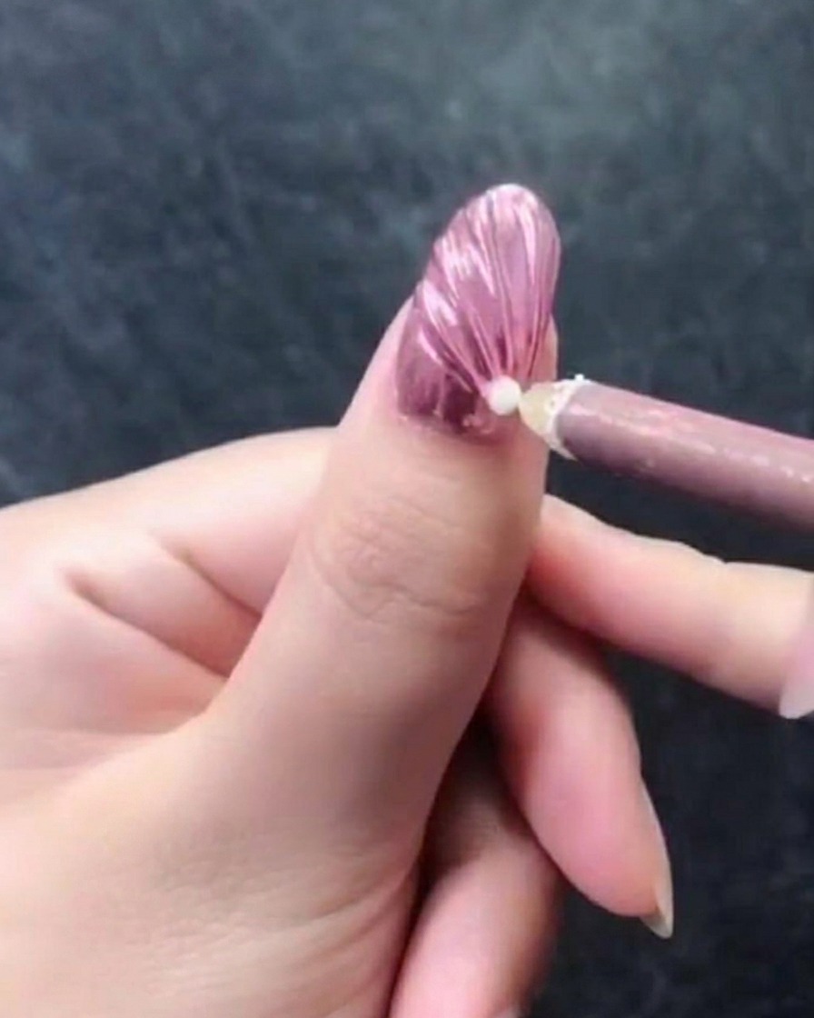 Nahaufnahme einer Hand mit einem rosa metallisch strukturierten Nagel, auf dem ein rosa Wachsstift eine einzelne weiße Perle in der Nähe der Nagelhaut platziert.