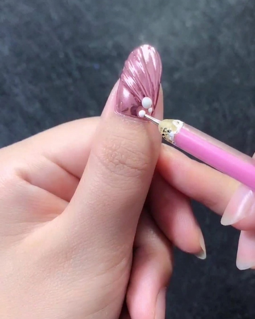 Nahaufnahme eines rosa metallisch strukturierten Nagels, bei dem ein Wachsstift winzige weiße Perlen direkt neben einer größeren weißen Perle an der Nagelhaut hinzufügt.