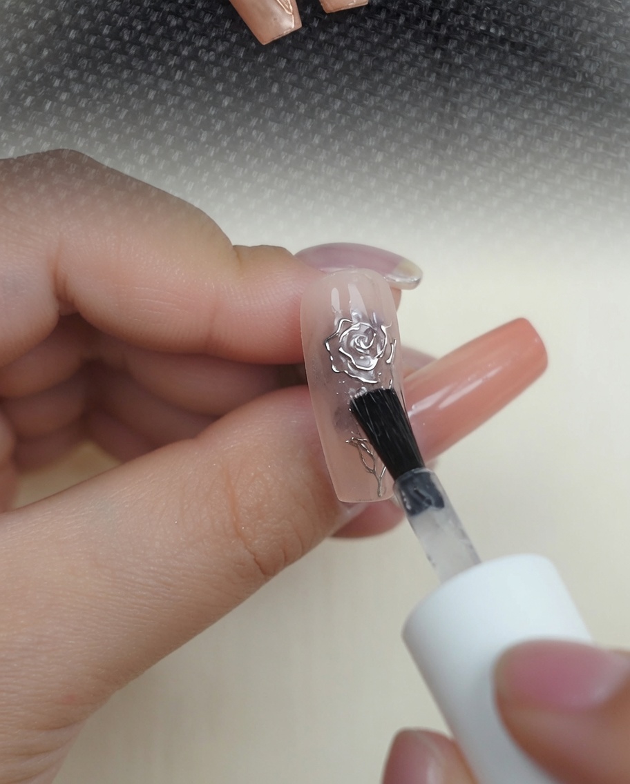 Ein Pinsel trägt klaren Hochglanz-Top-Coat über das metallische 3D-Rosendesign auf einem Nude-Nagel auf.