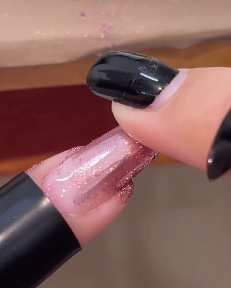 Eine Fingerspitze reibt roségoldenes Chrome-Puder über einen glänzenden Nagel.