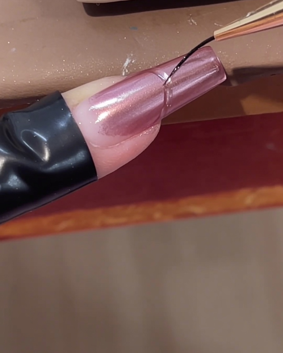 Ein feiner Linerpinsel fügt eine geschwungene, klare Aufbaugel-Linie nahe der Nagelhaut eines rosa Chrome-Nagels hinzu.