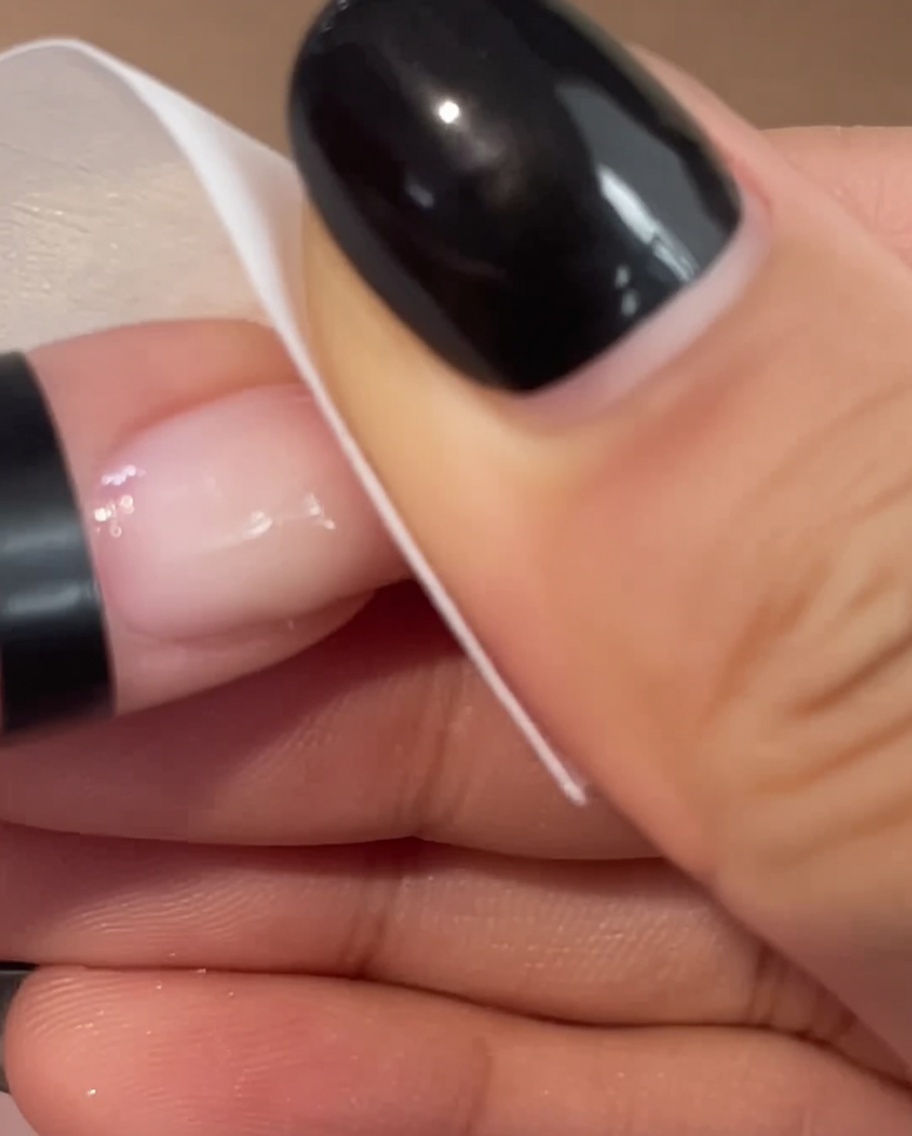 Nahaufnahme einer Hand, die mit einem weißen Tuch sanft die Oberfläche eines rosa manikürten Nagels reinigt.