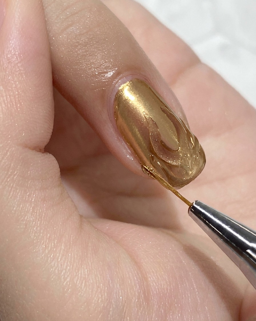 Nahaufnahme eines feinen Detailpinsels, der klares Hartgel innerhalb der Gold-Chrome-Flammenkontur auf einem zartrosa Fingernagel aufträgt.