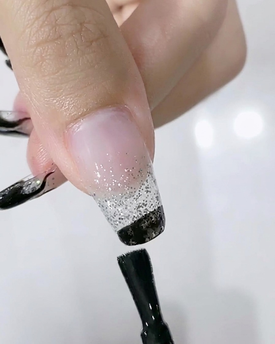 Ein Pinsel trägt schwarzes UV-Gel auf die freie Nagelkante über einem silbernen Glitzerverlauf auf.