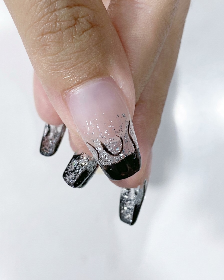 Eine Nahaufnahme eines Fingernagels, die eine fertige schwarze Flammenumrandung über einer silbernen Glitzerbasis zeigt.