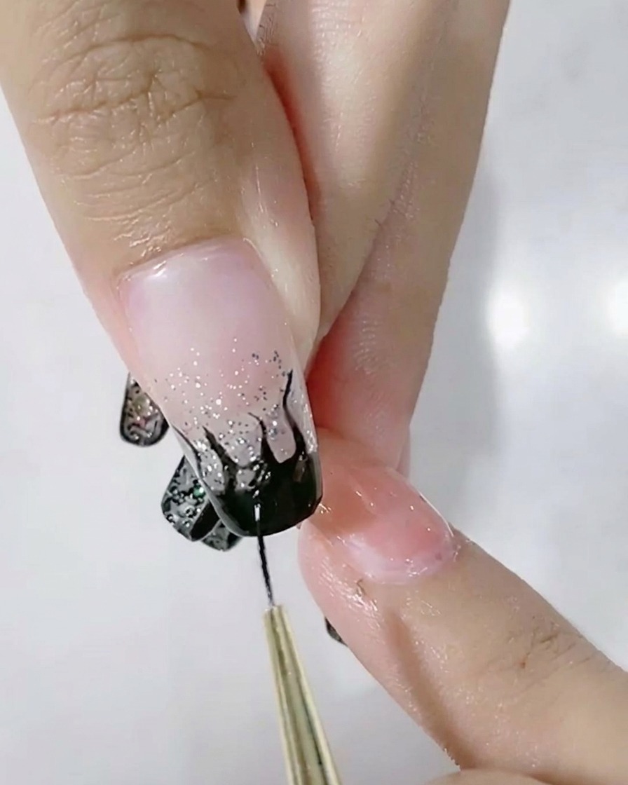 Ein feiner Pinsel füllt ein schwarzes Flammendesign auf einem Nagel mit Glitzerverlauf aus.