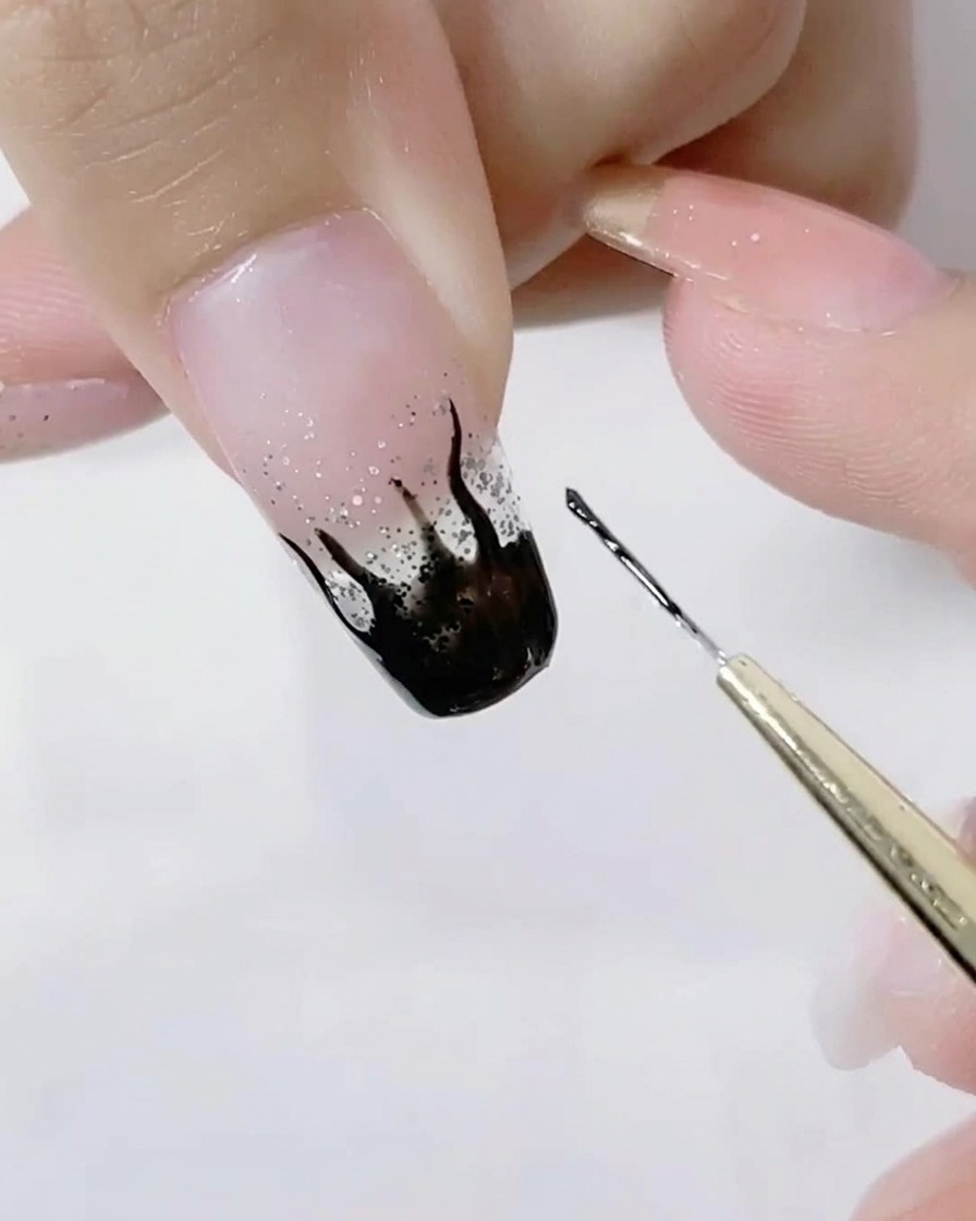 Nahaufnahme eines Pinsels, der die scharfen Spitzen der schwarzen Flammen auf einem Glitzernagel detailliert ausarbeitet.