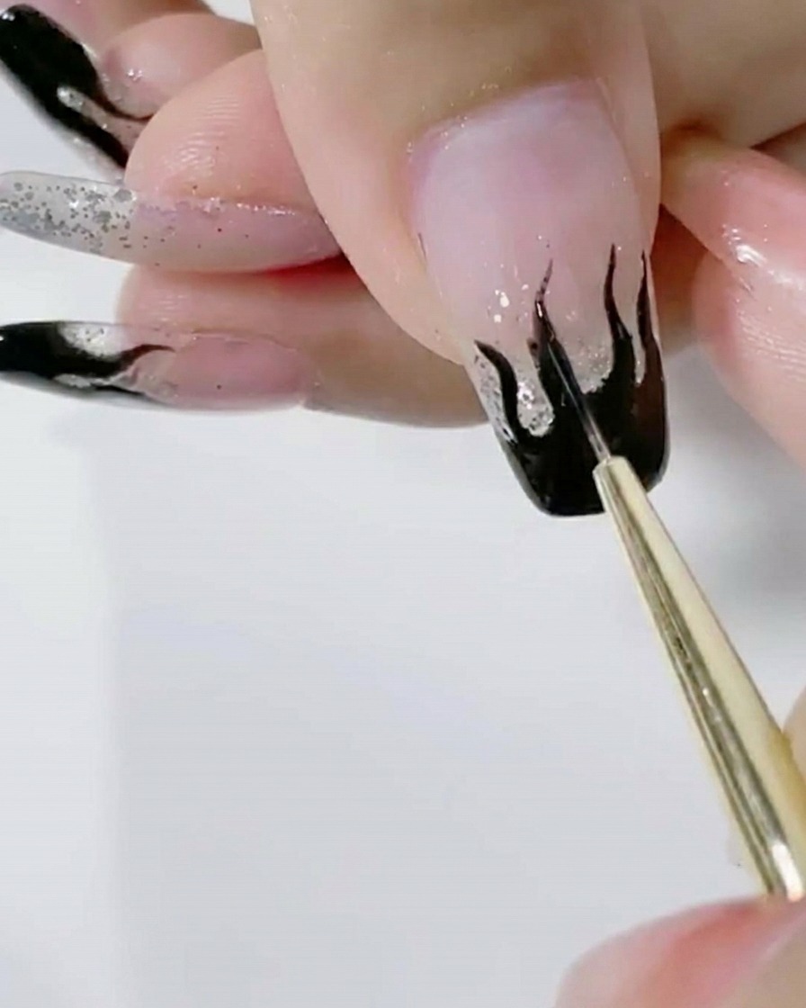 Ein Pinsel trägt eine zweite Schicht sattes schwarzes Gel auf das Flammendesign eines Glitzernagels auf.