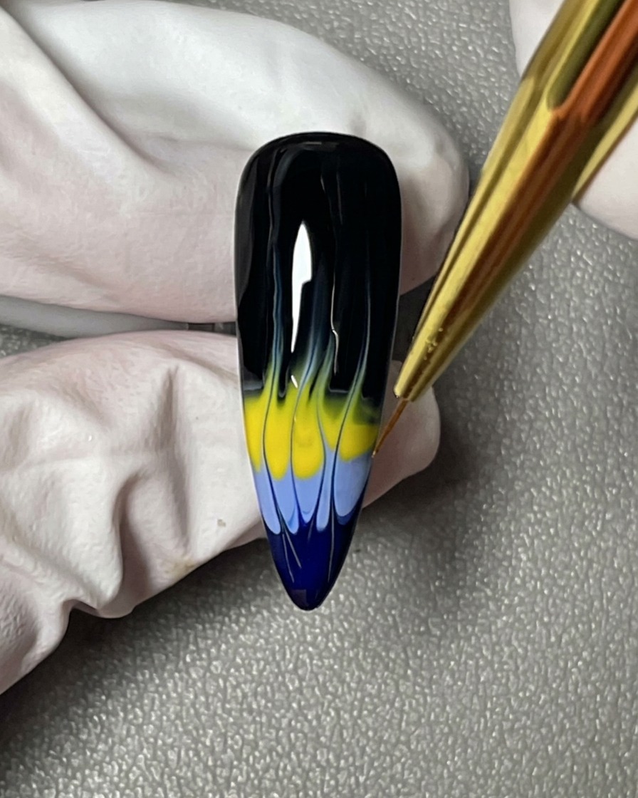 Nahaufnahme, die zeigt, wie ein Liner-Pinsel die scharfen, spitz zulaufenden Punkte des blau-gelben Flammendesigns auf einem schwarzen Nagel perfektioniert.