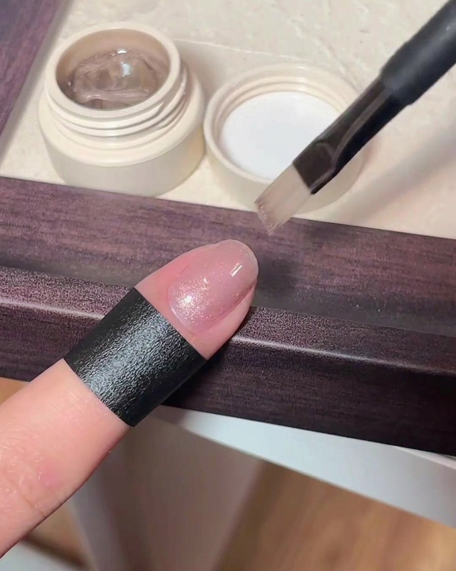 Nahaufnahme eines Pinsels, der ein transparentes, schimmerndes rosa Cat-Eye-Gel auf einen Naturnagel aufträgt.