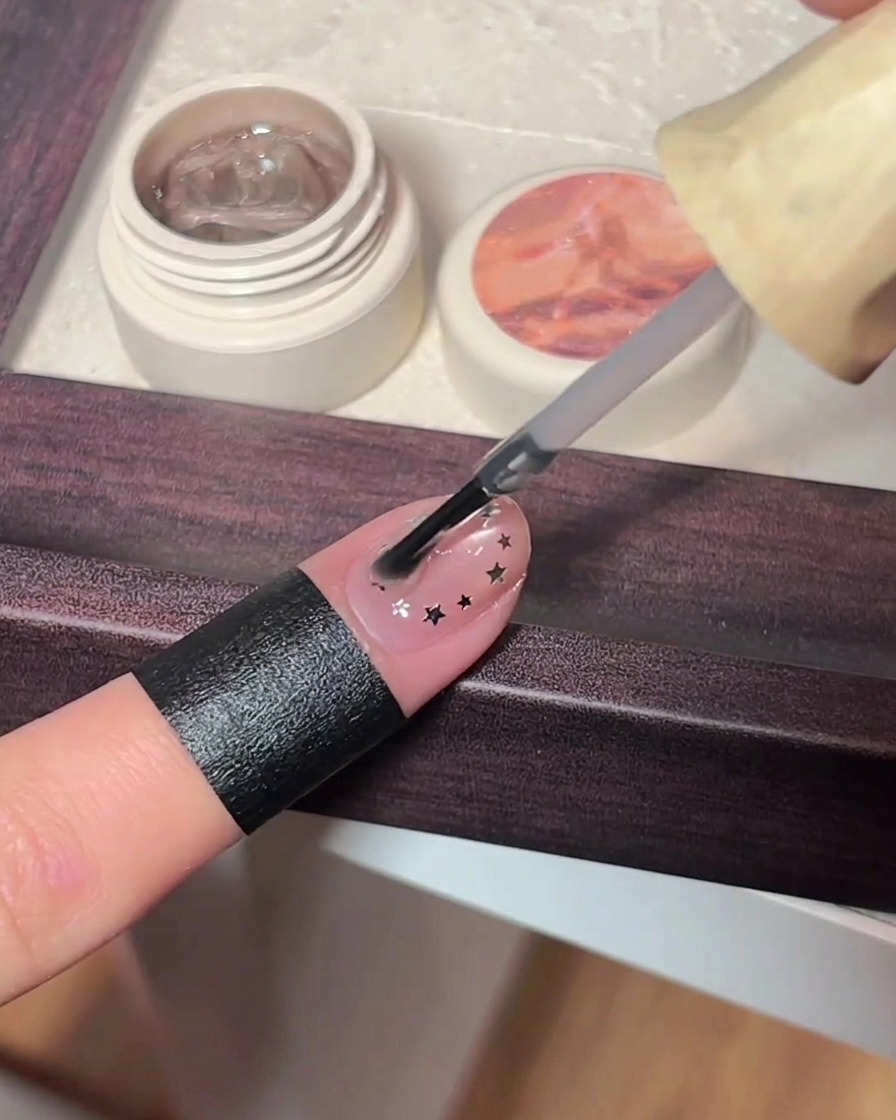 Ein Nagellackpinsel trägt eine dicke Schicht klaren Top-Coat über einen rosa Cat-Eye-Nagel auf, der mit winzigen metallischen Stern-Applikationen verziert ist.