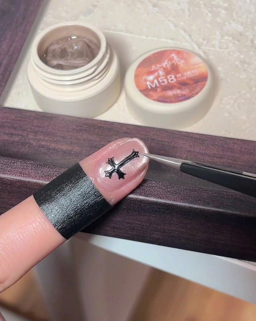 Eine Pinzette platziert einen kleinen, dunkelmetallischen Kreuz-Charm in der Mitte eines rosa Cat-Eye-Gelnagels.