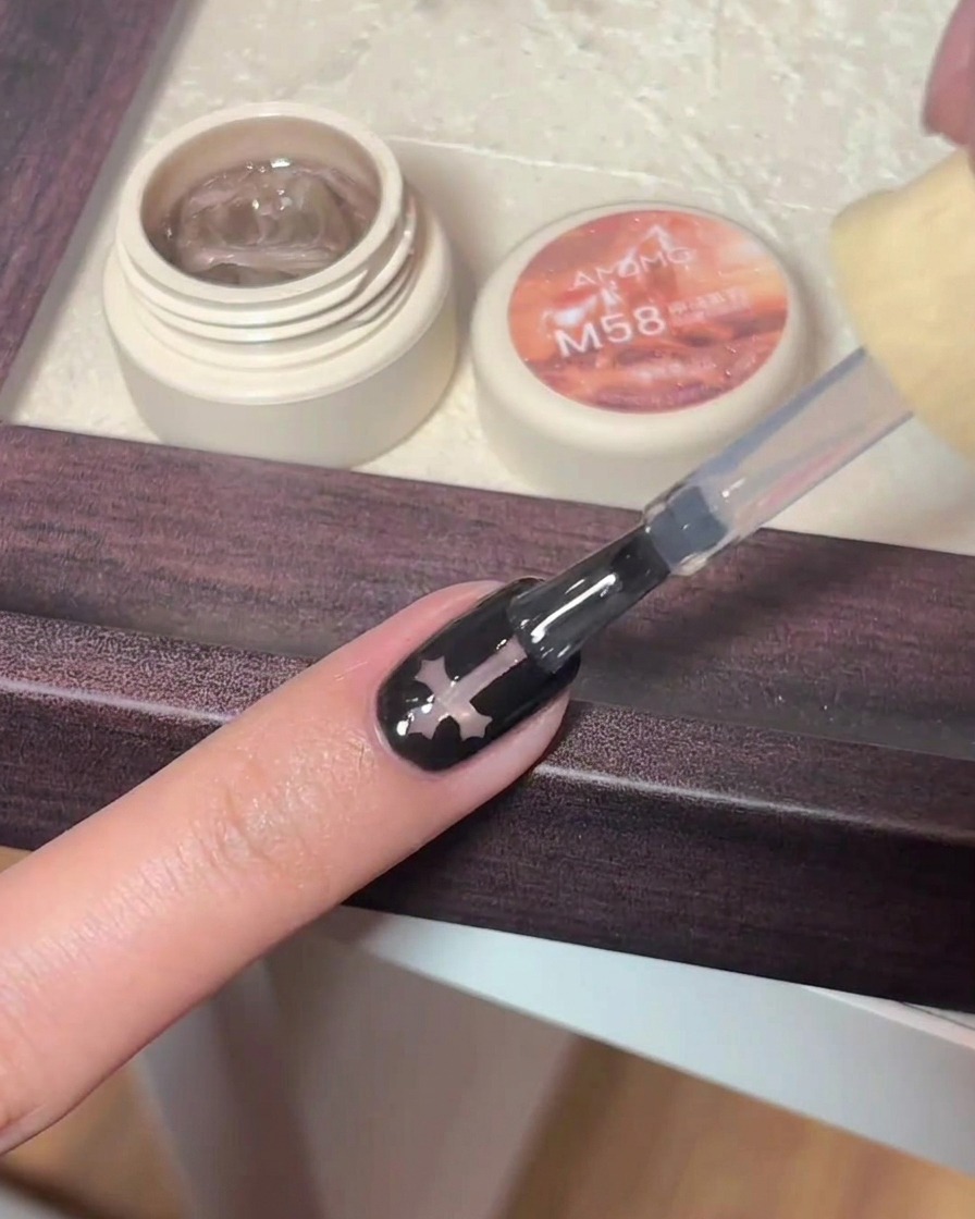 Ein Gel-Pinsel trägt glänzenden klaren Top-Coat über einen schwarzen Nagel mit Kreuz-Charm-Design auf.