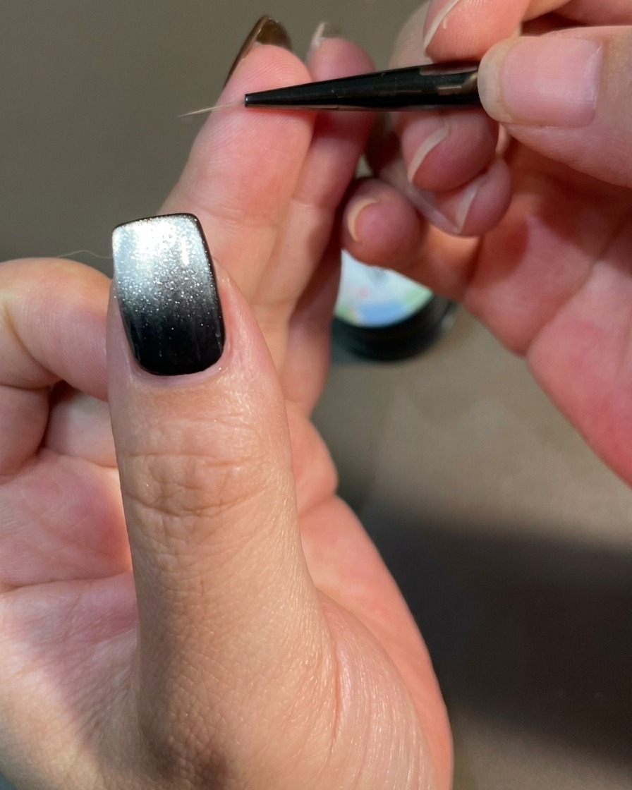 Nahaufnahme eines Nagels, der mit einem feinen Liner-Pinsel verziert wird, wobei geschwungene, flammenartige Linien auf eine schwarze magnetische Gel-Basis gezeichnet werden.