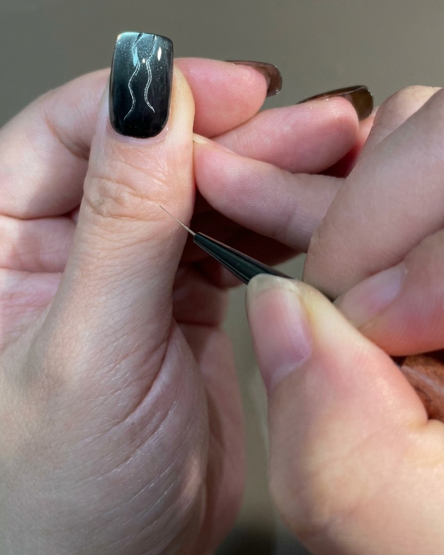 Nahaufnahme eines Nagels, auf dem mit einem Liner-Pinsel mehrere geschwungene Linien unterschiedlicher Länge über eine magnetische Gel-Basis gezeichnet werden.